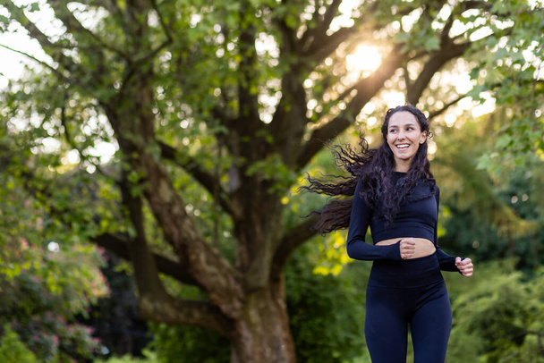 Young motivated and joyful woman jogging in the morning in a public park among trees, Hispanic woman with curly hair, slim in a tracksuit. - Foto, Imagem