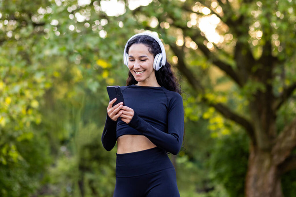 Young beautiful Latin American woman walks in the park during active physical exercises, woman uses an application for listening to audio books and music on phone, smiling sportswoman in headphones. - Fotografie, Obrázek