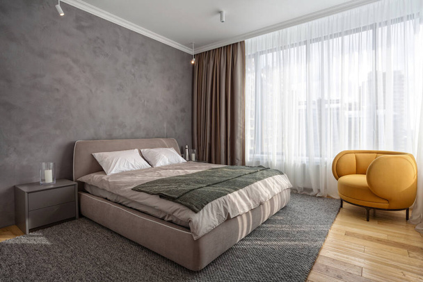 Light modern bedroom with comfortable furniture on natural wooden floor. Double bed on woven rug, close to bedside table and classic orange armchair. Concept of minimalist interior design in apartment - Photo, Image