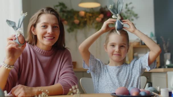 Portret van mooie moeder en schattig dochtertje glimlachend op camera terwijl poseren met versierde paaseieren aan tafel in de keuken - Video