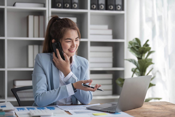Portret młodej biznesmenki siedzącej w biurze przed laptopem i rozmawiającej przez telefon komórkowy w biurze. - Zdjęcie, obraz