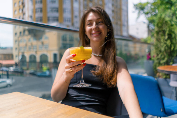 egy fiatal mosolygós lány koktéllal a kezében ül egy asztalnál egy nyári teraszon, kilátással a városra a bárban. - Fotó, kép