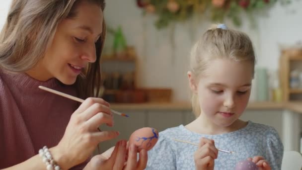 Close up tilt down shot of beautiful mother and little daughter using brushes for decorating Easter eggs with paint while preparing home for holiday - Footage, Video