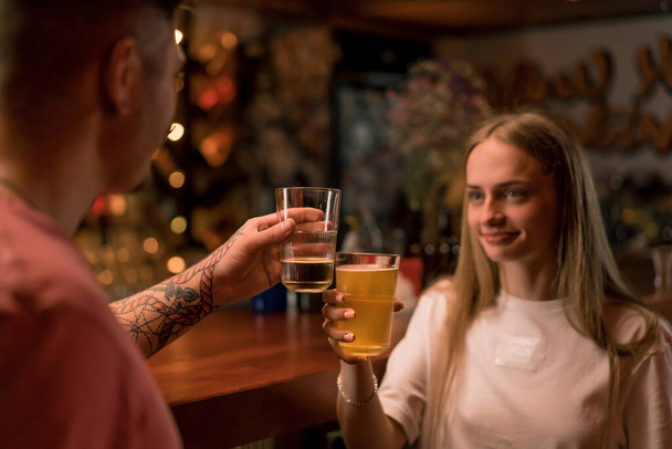 Elinde bir bardak birayla gülümseyen genç bir kız bardaki bir adamla sohbet ediyor. - Fotoğraf, Görsel