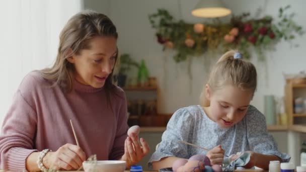 Mediana toma de madre feliz y pequeña hija decorando huevos teñidos y haciendo artesanía de papel mientras se prepara para la Pascua en casa - Metraje, vídeo