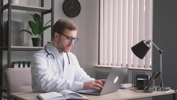 Ένας νεαρός ελκυστικός νευρολόγος γιατρός κάθεται στο γραφείο και εργάζεται στον υπολογιστή. Ένας νεαρός ειδικευόμενος δουλεύει σε ένα βιβλιάριο δικτύου. Κάθετη βίντεο - Πλάνα, βίντεο