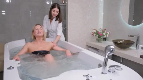 Relaxed, beautiful woman lying in a hot tub in a spa salon. A beautician gives a health massage to a client. - Кадры, видео