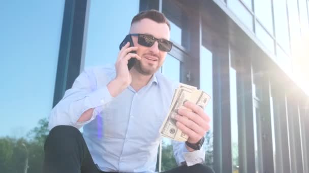Een jonge baas met dollars in zijn handen praat op een smartphone. Bedrijfsmanager concept. Verticale video - Video