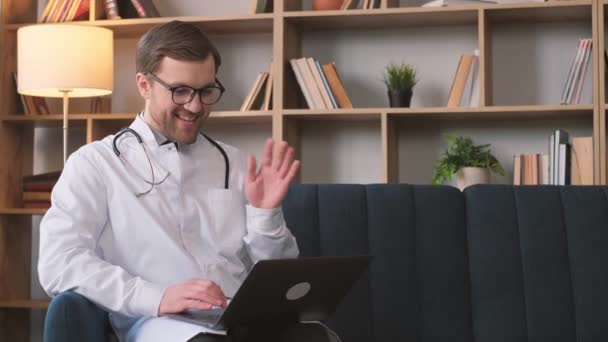 Un joven médico barbudo con un uniforme blanco y gafas está sentado en una habitación de residentes y hablando con alguien en Internet. Especialista médico joven - Metraje, vídeo