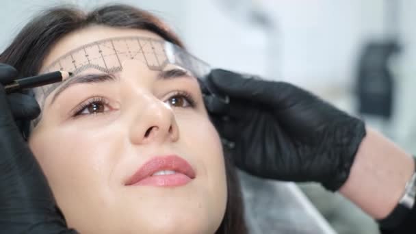 A close-up beautician corrects the shape of the eyebrows with a special ruler. Beautiful girl in a beauty salon - Πλάνα, βίντεο