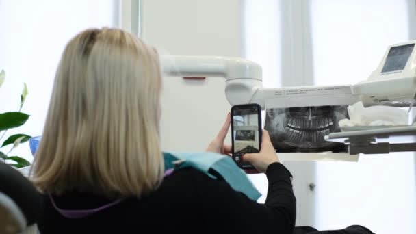 Una paciente rubia toma una radiografía de sus dientes en un teléfono inteligente mientras está sentada en una silla dental. Un paciente joven en un consultorio dental - Imágenes, Vídeo