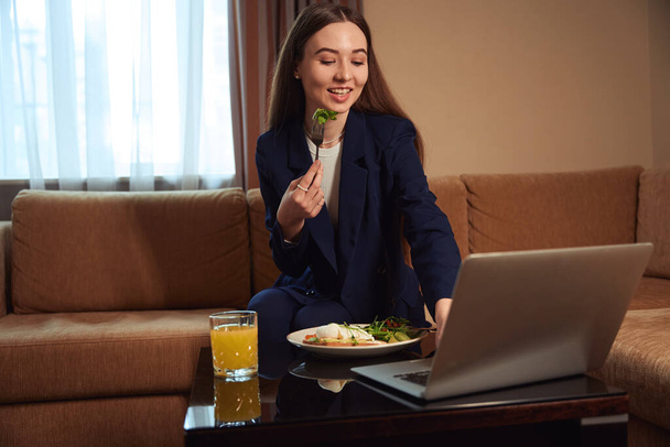 Улыбающаяся женщина в деловом костюме сидит на диване и завтракает, смотрит кино в номере отеля - Фото, изображение