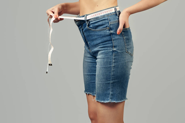 Koncepcja odchudzania. Dziewczyna trzyma taśmę pomiarową owiniętą wokół talii zamiast paska na jeansowych spodenkach. Zbliżenie. - Zdjęcie, obraz