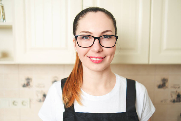 Ein Mädchen in Schürze und Brille blickt lächelnd in die Kamera, während sie in der Küche steht. Hausfrauenkonzept. - Foto, Bild