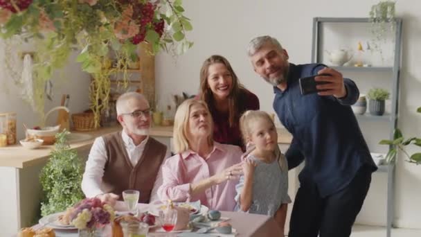 Brede opname van gelukkige familie zwaaien en glimlachen op smartphone camera terwijl het nemen van selfie of video bellen op Pasen diner thuis - Video