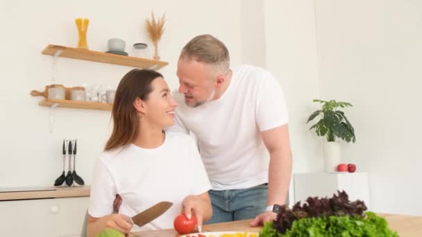  Egy fiatal nő zöldségeket aprít, hogy egészséges salátát készítsen, miközben a férje megöleli a konyhában. Függőleges videó - Felvétel, videó