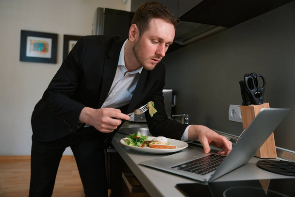 Jovem do sexo masculino em terno de negócios em pé perto da mesa e comer, digitando no laptop no quarto do hotel - Foto, Imagem