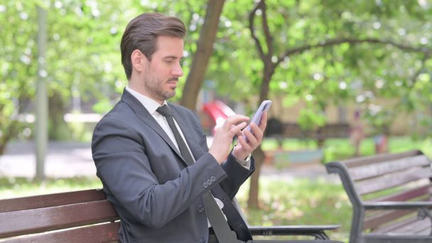Молодой бизнесмен просматривает интернет-смартфон, сидя на скамейке запасных - Фото, изображение