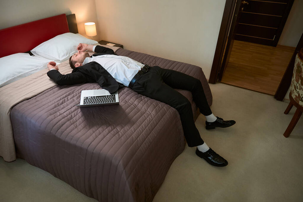 Młody mężczyzna w garniturze biznesowym odpoczywający na łóżku przy laptopie w pokoju hotelowym - Zdjęcie, obraz