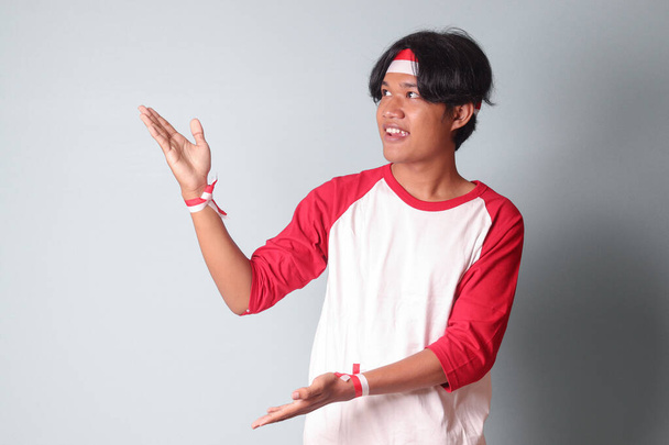 Portrait of attractive Asian man in t-shirt with red and white ribbon on head, pointing at something with finger. Isolated image on gray background - Photo, Image