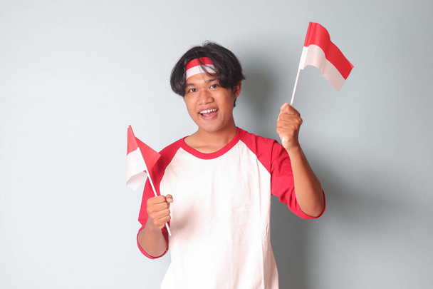 Portrait of attractive Asian man in t-shirt with red and white ribbon on head, holding indonesia flag while raising his fist, celebrating success. Isolated image on gray background - Photo, Image