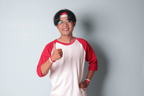 Портрет привлекательного азиатского мужчины в футболке с красной и белой лентой на голове, показывающего хороший жест рукой с большим пальцем вверх. Изолированное изображение на сером фоне - Фото, изображение