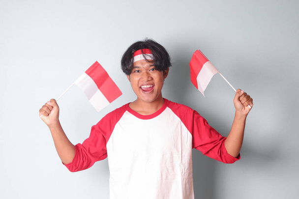 Портрет привабливого азіатського чоловіка в футболці з червоно-білою стрічкою на голові, тримаючи прапор Індонезії, піднімаючи кулак, святкуючи успіх. Ізольоване зображення на сірому фоні - Фото, зображення