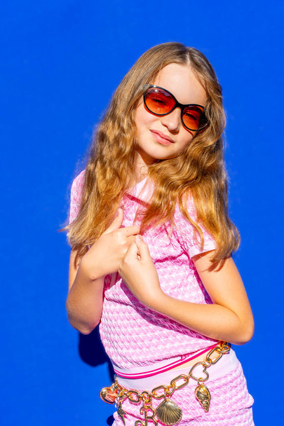 Attractive model Barbie style girl wearing sunglasses, pink tee-shirt and shorts with long hair on blue backdrop, vintage fashion concept. Hight quality photo. - Foto, Bild