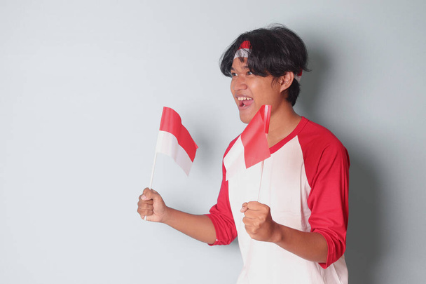 Portrait of attractive Asian man in t-shirt with red and white ribbon on head, holding indonesia flag while raising his fist, celebrating success. Isolated image on gray background - Photo, Image