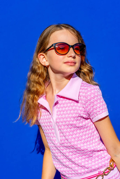 Ελκυστικό μοντέλο Barbie στυλ κορίτσι φορώντας γυαλιά ηλίου, ροζ μπλουζάκι και σορτς με μακριά μαλλιά σε μπλε φόντο, vintage έννοια της μόδας. Φωτογραφία υψηλής ποιότητας. - Φωτογραφία, εικόνα