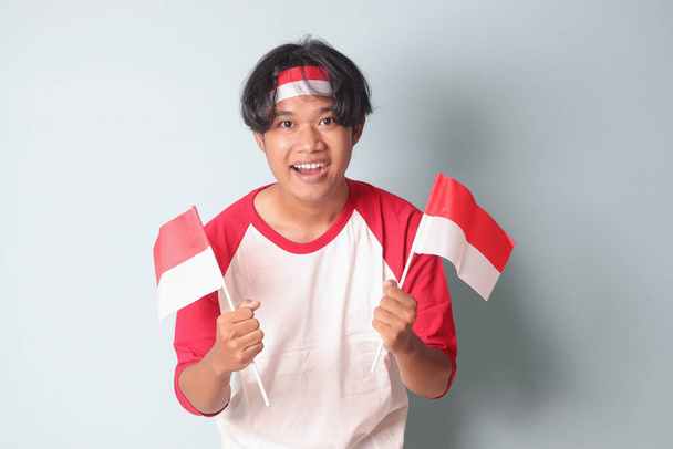 赤と白のリボンを頭に赤と白のリボンで着た魅力的なアジア人男性の肖像画は,彼の拳を上げながらインドネシアの旗を保持し,成功を祝います. 灰色の背景に隔離された画像 - 写真・画像