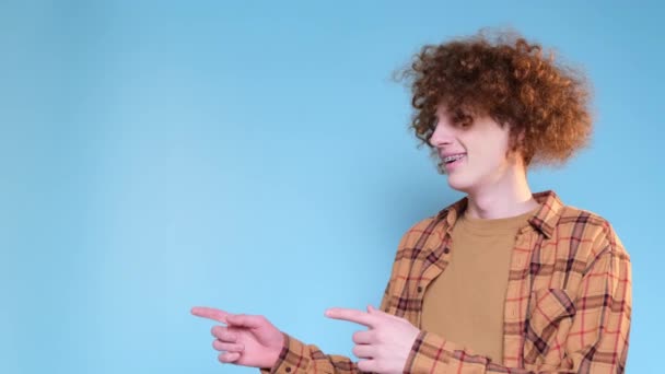 Red-haired positive young man in shirt pointing hands at someone while standing against blue background. Hand gestures. A student with a strange hairstyle. - Footage, Video