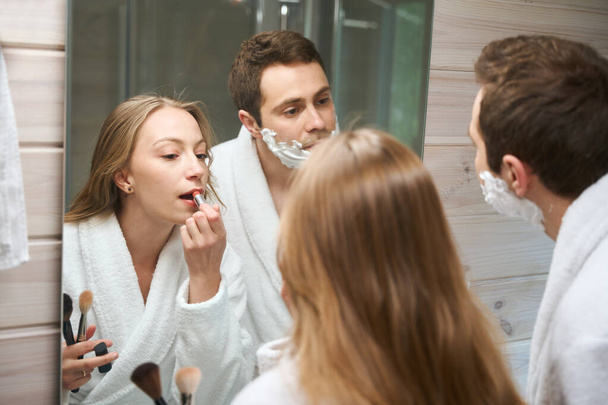 Young couple in the bathroom, woman paints her lips, man looks in the mirror - Photo, Image