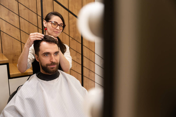 Artisanat travaille avec un client dans un salon de coiffure moderne, un homme en cape de protection - Photo, image