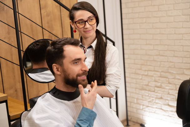 Giovane barbiere dimostra la sua acconciatura dal retro al cliente allo specchio, il cliente sorride - Foto, immagini