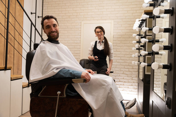 Un client barbu est assis dans une chaise de coiffure, à côté d'un coiffeur féminin avec des outils de travail - Photo, image
