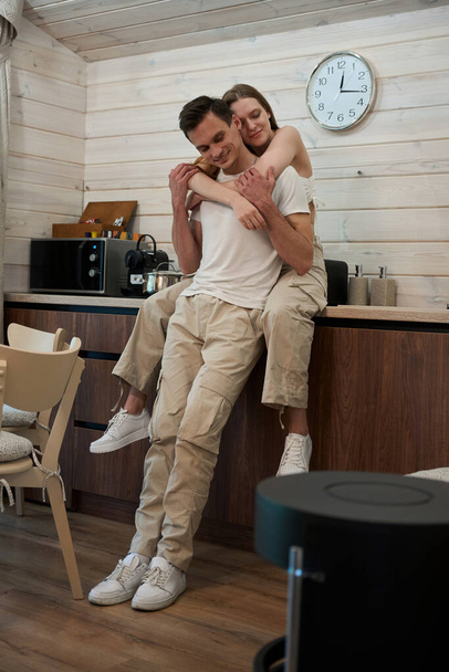 Donna abbracciare un uomo in piedi accanto a lei mentre seduto su un tavolo in cucina - Foto, immagini
