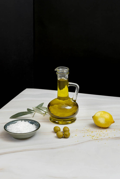 A beautiful still life with a glass container filled with virgin olive oil, lemon zest, salt flakes, olives and olive leaves - Valokuva, kuva