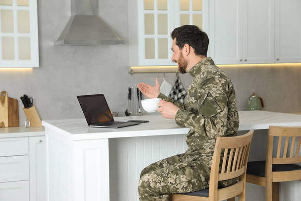 Soldier with cup of drink using video chat on laptop at white marble table in kitchen. Military service - Fotó, kép