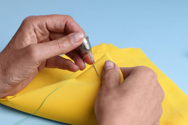 Woman sewing on yellow cloth with thimble and needle against light blue background, closeup - Fotó, kép