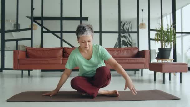 Стара жінка робить вправи на йогу вдома. Зріла жінка має медитацію. вона сідає на ліжко, дивиться у вікно і радий, що в неї є час відпочити. Високоякісні 4k кадри - Кадри, відео