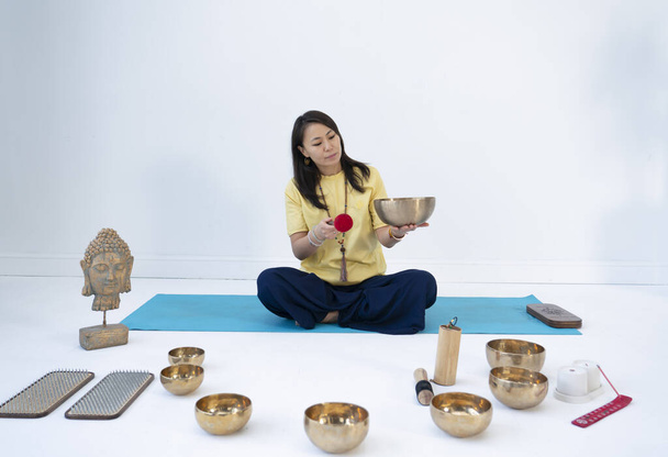 A young Asian woman sits in a lotus position and taps on a Tibetan singing bowl. High quality photo - Photo, Image