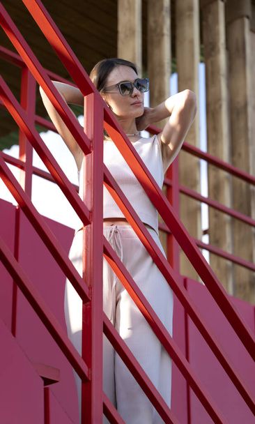  Zomer stedelijk portret van een jonge brunette in een zonnebril. Een vrouw staat op een toren met rode leuningen. Hoge kwaliteit foto - Foto, afbeelding