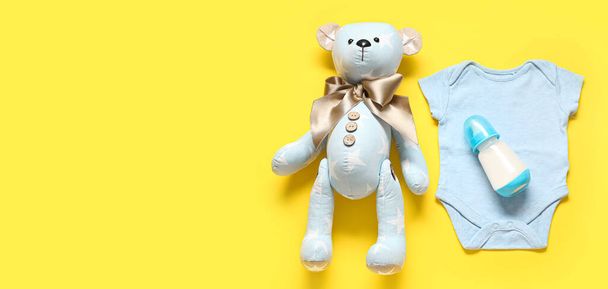 黄色い背景の赤ちゃんのためのミルクのボトルが付いている熊のおもちゃおよびボディスーツ. デザインのためのバナー - 写真・画像