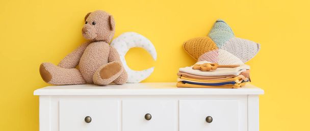 Stapel Babykleidung mit Spielzeug auf Tisch neben gelber Wand - Foto, Bild