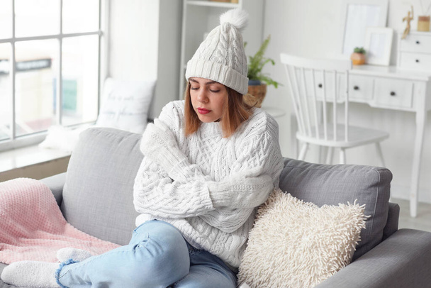 Frozen young woman in winter clothes sitting on sofa at home - Foto, imagen