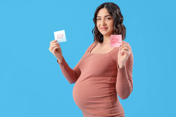 Jonge zwangere vrouw met woorden BOY en GIRL op papier tegen blauwe achtergrond - Foto, afbeelding