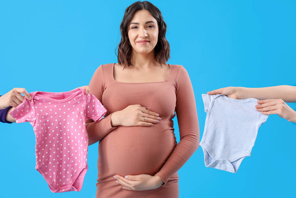Young pregnant woman and female hands with baby bodysuits on blue background - Foto, imagen