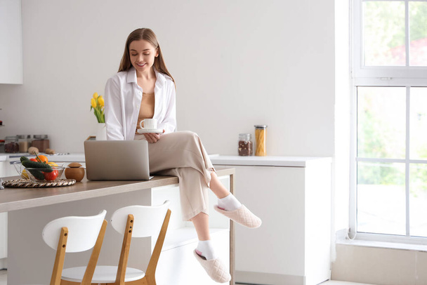 Happy young woman holding cup of coffee and sitting on table with modern laptop in light kitchen - Photo, Image