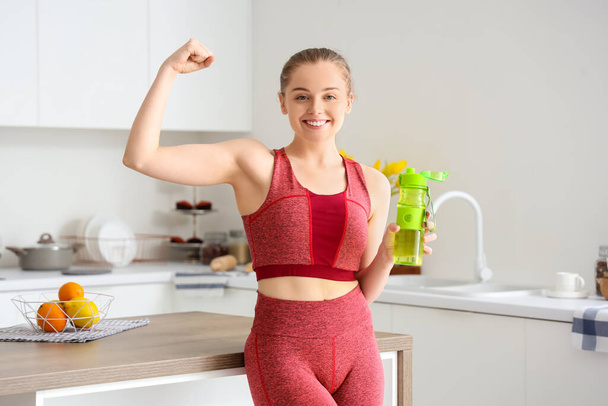 Спортивная молодая женщина с бутылкой воды разминает мышцы рук на светлой кухне - Фото, изображение
